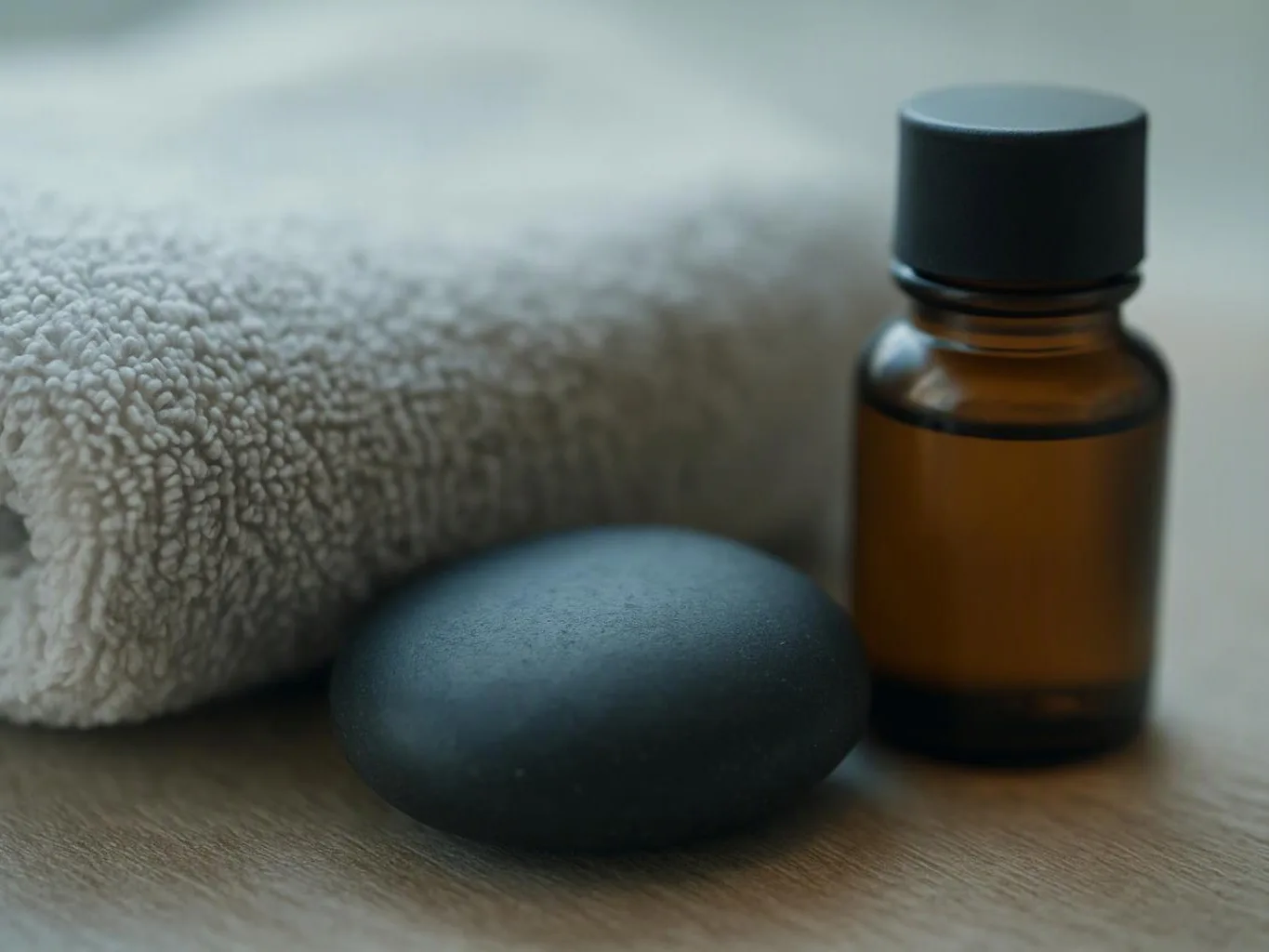A towel, black massage stone, and amber bottle sit on a wooden surface